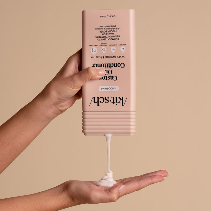 Hand holding a pink bottle of conditioner with visible cream being dispensed onto a palm against a beige background