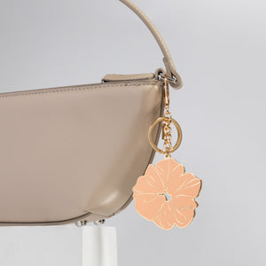 Beige handbag with a metal enamel keychain depicting the kitsch Terracotta Scrunchie with gold outline, design, and hardware on a light gray background
