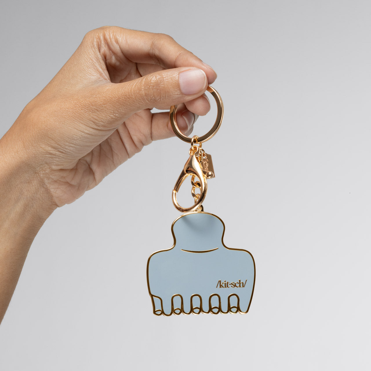 Hand holding a Metal enamel keychain of the kitsch Haze Blue Cloud Clip with a gold outline and hardware against a light gray background
