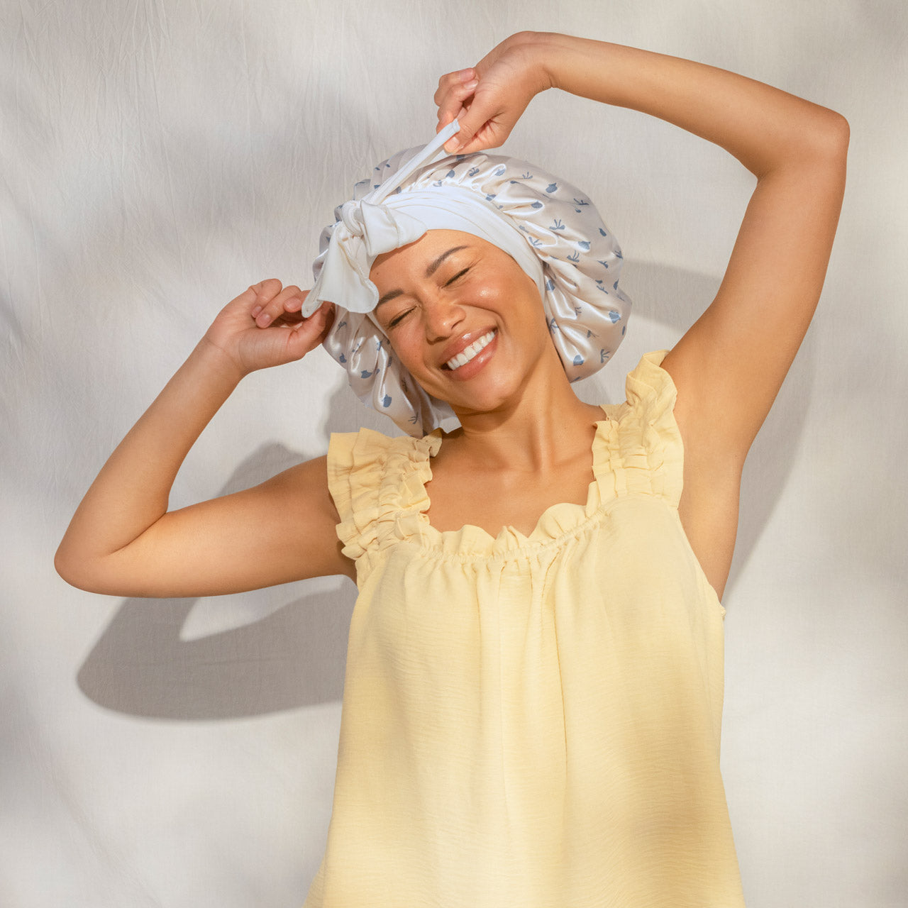 Smiling female model in yellow ruffled top wearing KITSCH Pearlized Cream & Coastal Blue Print Oversized Satin Bonnet — arms raised, joyful lifestyle portrait shot — Adjustable, double-layered satin — For preserving your hair overnight