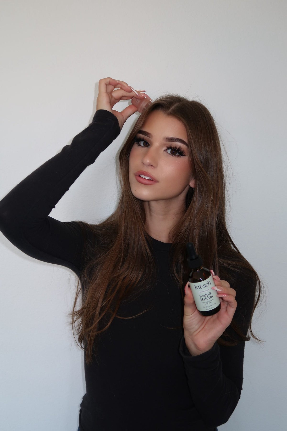 Brooke Monk running a hand through her long brown hair, holding the Kitsch Rosemary Scalp & Hair Oil